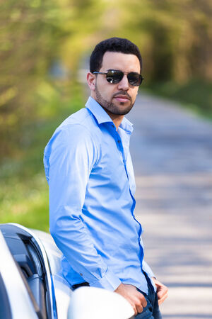 Young latin american driver seated on the side of his new carの写真素材