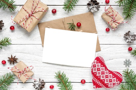 Christmas greeting card in frame made of fir branches, red berries, knitted heart, gift boxes and pine cones on white wooden table. Christmas background. Flat lay. top view with copy spaceの写真素材