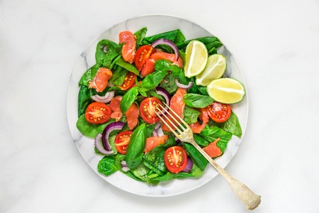 Salmon salad with spinach, cherry tomatoes, red onion, lemon and basil in a plate with fork over white marble background. healthy food concept. top viewの写真素材