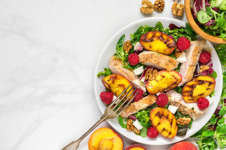 vitamin salad with grilled chicken and peach, feta cheese, raspberries and walnuts in a plate with fork over white marble table. healthy food. top view with copy spaceの写真素材