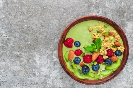 bowl of green matcha tea smoothie with fresh berries, fruits, granola, nuts and seeds for healthy...