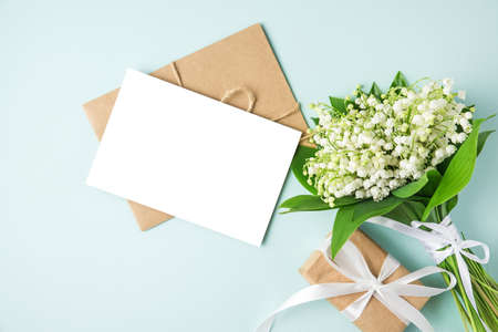 blank greeting card with lily of the valley flowers bouquet and gift box on blue background. mock up. flat lay. top view. wedding invitationの写真素材