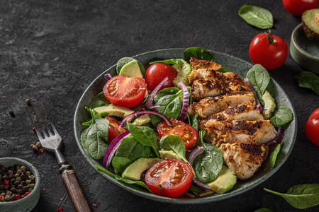 Grilled chicken breast fillet and spinach salad with avocado, tomatoes and sesame seeds in a plate on dark background with fork. close upの写真素材