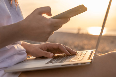 Close up of woman working with laptop and mobile phone on the beach by the sea during sunset. Female freelancer work online using smartphone and computer from a distanceの写真素材