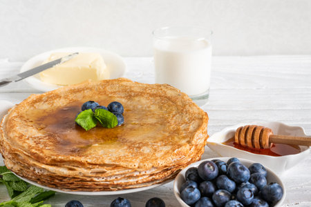 Stack of thin pancakes with blueberries, mint and honey. Tasty breakfast with french crepes, milk and butter on white wooden table. Delicious foodの写真素材