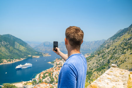 Man tourist taking photo on smartphone standing on Lovcen mountain above Kotor bay and old town, Montenegro. Male traveler taking picture on mobile phone of beautiful view of Adriatic fjordの写真素材