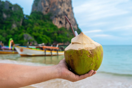 Male hand holding fresh coconut drink on tropical sandy Railay beach with cliffs and traditional Thai boats at Krabi province, Thailand. Man holds refreshment summer drink with a strawの写真素材