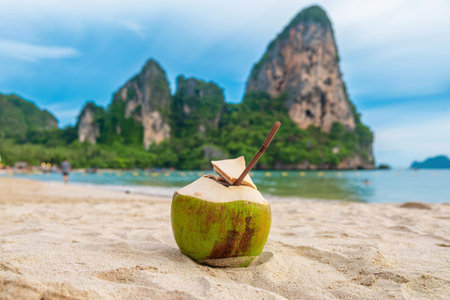 Fresh coconut drink with a straw on sandy tropical Railay beach, with lush green cliffs and clear blue water in the background, Krabi province, Thailand. Refreshment summer drinkの写真素材