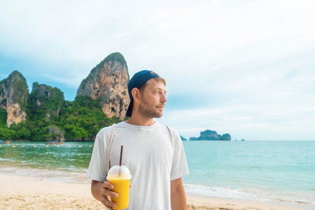 Cheerful man tourist hold yellow mango smoothie in plastic cup with a straw on tropical sandy Railay beach with cliffs at Krabi province, Thailand. Happy male with refreshment summer drinkの写真素材