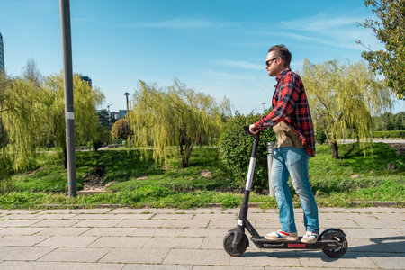 Side view of man riding e-scooter on pavement surface in city public park at sunny summer day. Male driving electric scooter outdoors. Eco-friendly city transport and travel. Sustainable lifestyleの写真素材
