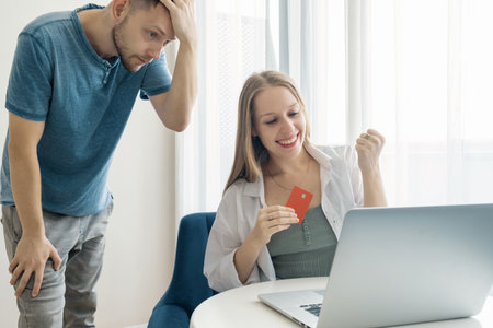 Happy woman celebrates successful online shopping with credit card while her male partner looks...