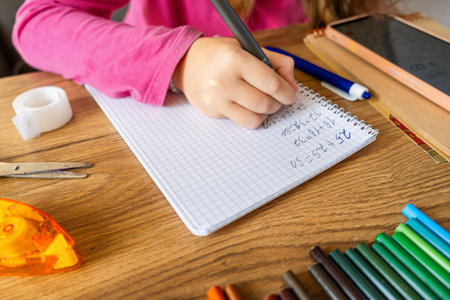 Child doing math exercises in notebook using smartphone with stationery on wooden table. Girl hand writing mathematical equations as school homework. Learning, education using digital tools concept.の写真素材