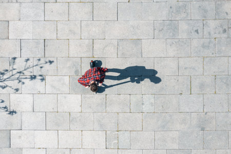 Top view of man riding electric scooter on paved surface casting shadow in the city at sunny summer day. Overhead view of male driving e-scooter outdoors. Artistic and minimalictic shot from droneの写真素材