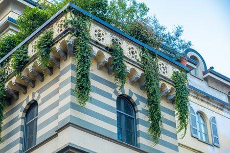 Historic building facade with striped pattern and hanging plants cascade from rooftop, combining traditional architecture with natural greenery, Milan, Italy. Eco friendly urban environmentの写真素材