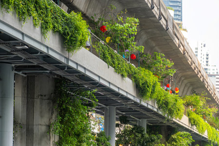 Elevated walkway with lush greenery, climbing plants, and colorful lanterns under train tracks in...