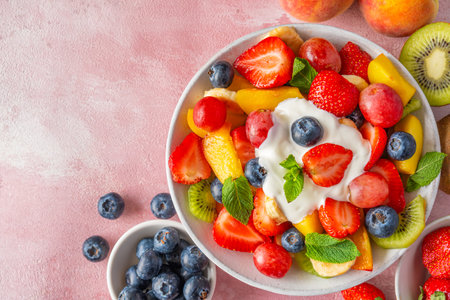 Summer colorful fruit salad in a bowl creamy yogurt on pink background. Top view with copy space. Healthy vitamin food for breakfast. Mixed strawberries, grapes, banana, kiwi, blueberries, peach, mintの写真素材