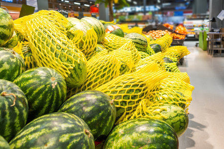 Fresh whole watermelons stacked in supermarket display wrapped in yellow protective mesh for sale. Freshness, abundance in grocery store in Thailand. Food retailの写真素材