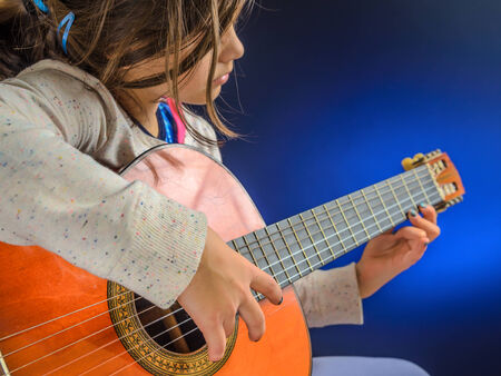 Little girl plays red shiny classical guitar, blue light, performance, concert, focus on right hand.