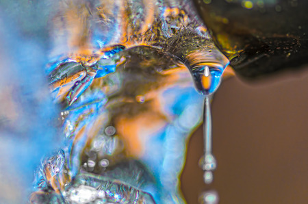 Drop of melting ice water at the drainpipe, with reflections of blue sky and yellow sun rays passing through. Macro with ice structure seen through the drop of water.の写真素材