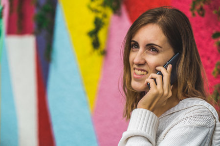 A woman's beaming smile brightens a phone conversation against an abstract backdrop of vibrant geometric patterns, seamlessly blending modern artistry with genuine joyの写真素材