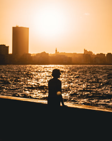 A woman stands upon Havana's wave breaker, captivated by the sunset's allure. The rhythmic crashing of waves harmonizes with the vibrant colors, painting a breathtaking scene of serenity and wonderの写真素材