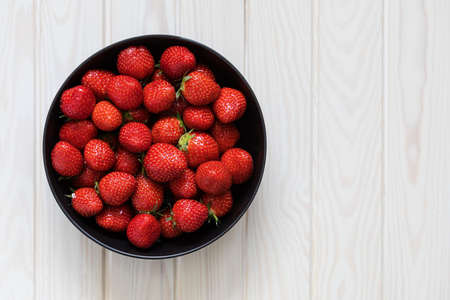 Black bowl with fresh strawberry berries on a wooden light background