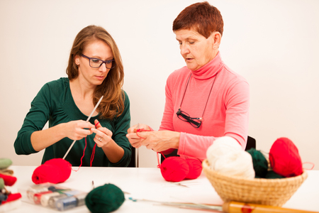 Women knitting with red wool. Eldery woman transfering her knowledge of knitting on a younger woman on handcraft workshop.の写真素材