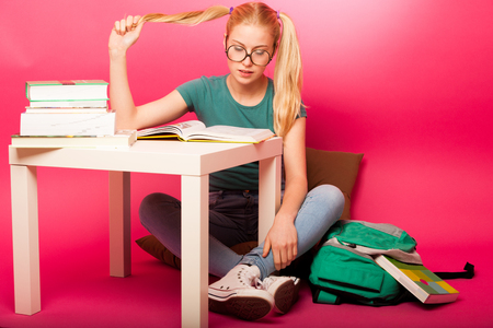 Playful schoolgirl with big eyeglasses concentrated in reading book sitting on floor behind the small table. Playing with one hair tail.の写真素材