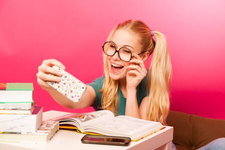 Curious, naughty, playful schoolgirl with big eyeglasses doing selfie with smartphone while study.