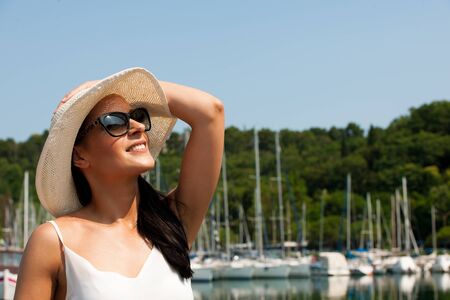 young woman with hat summer dress and sunglasses walks pier of marina on hot summer dayの写真素材