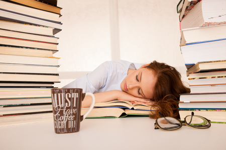 attractive beautiful tired student sleeps on pile of books with a coffee mug on a tableの写真素材