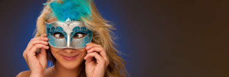 beauty portrait of Beautiful young blonde woman with carnival mask in hands over blue backgroundの写真素材