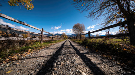 A stunning view of a gravel pathway meandering through the countryside, flanked by a wooden fence under a radiant blue sky, perfect for nature lovers.の素材