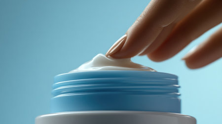 A close-up view of a hand gently touching a creamy moisturizer on a container, showcasing skincare in a soothing blue background. This image highlights beauty and self-care.の素材