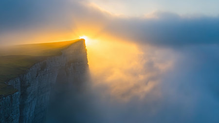 A breathtaking sunrise illuminates a rugged mountain cliff, casting golden light through swirling fog, creating a serene and picturesque landscape.の素材