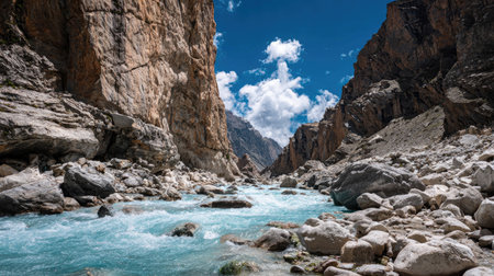 Breathtaking view of a rocky canyon with a clear turquoise river flowing between large boulders, under a vivid blue sky adorned with fluffy white clouds.の素材