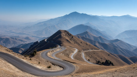 A breathtaking view of a winding mountain road snaking through rugged terrain, set against a backdrop of distant mountains under a clear blue sky, perfect for travel enthusiasts.の素材