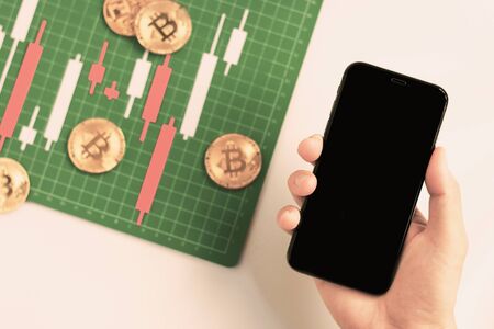 Top view hand holding smartphone with Candlestick chart make from color paper white and red on Green board with grid lines, investment, trading, stock, finance conceptの写真素材