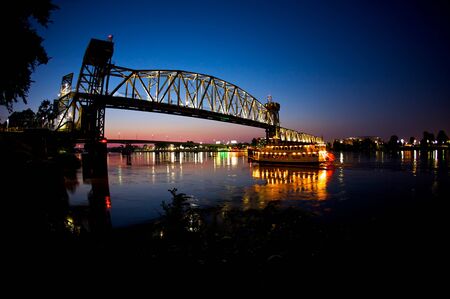A riverboat passes under a bridge at sunsetの写真素材