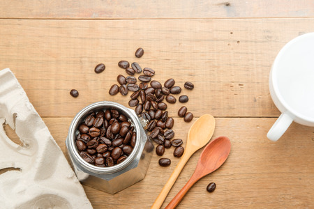 coffee beans in a moka pot close to couple of wood spoon and a take home tray of coffee cups and white coffee cups on wooden tableの写真素材
