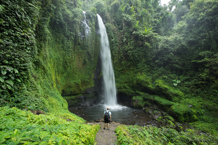 Man standing by huge tropical waterfall surrounded by lush green Rain forest vegetation and Jungle in Lombok, Indonesia