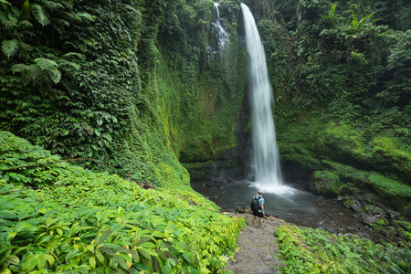 Man standing by huge tropical waterfall surrounded by lush green Rain forest vegetation and Jungle in Lombok, Indonesiaの写真素材