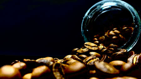 Whole raw coffee beans poured out of a jar on a table on a black backgroundの写真素材