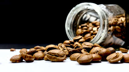 Whole raw coffee beans poured out of a jar on a table on a black background