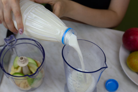 Girl hands pour milk from bottle into measuring cup to add to fruit in blender. Recipe for simple healthy fruit milkshake at home, ingredients for vitamin cocktailの写真素材
