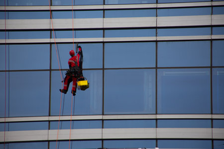Industrial climber in red overalls cleans the glass windows of skyscraper with office space, hard work at height for cleanliness of building exteriorの写真素材