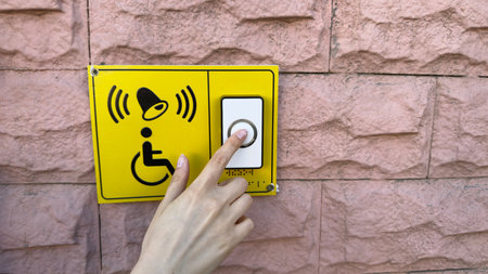 Closeup of female hand pressing accessibility call button for wheelchair users on yellow panel fixed to pink wall, symbolizing care, assistance and inclusive environmentの写真素材