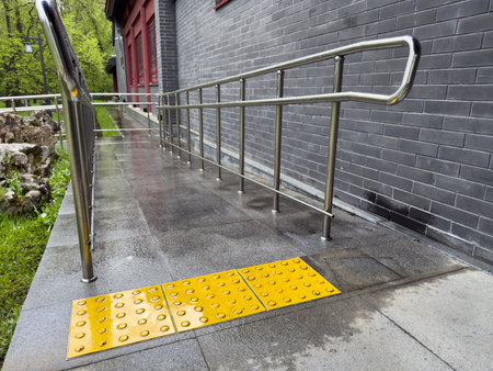 Tactile paving tiles with raised dots installed in front of accessibility ramp with metal handrails, providing guidance and safety for visually impaired individuals near building with brick wallの写真素材