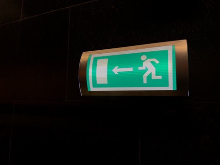 Illuminated green emergency exit sign on dark wall with arrow pointing left, symbolizing clear safety guidance and effective evacuation routes in case of fire or other critical situationsの写真素材
