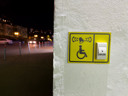 Accessible call button for people with disabilities mounted on textured wall in urban area at night. Clear signage with tactile surface and visual symbol for inclusive designの写真素材